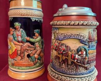 German beer steins with relief decoration, one featuring the Oktoberfest motif. Both have pewter lids with embossing and are marked on their base. The tallest stein measures 9 inches high.