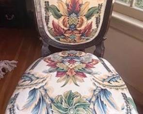 Carved wood chair with ornate crest, floral-patterned upholstery on seat and back, and decorative trim. Features turned front legs and casters. Back and seat display coordinated patterned fabric with vivid botanical motifs.