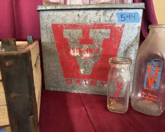 Clear glass milk bottles featuring Virginia Dairy logos in a wooden carrier marked "VIRGINIA DAIRY INC." and a metal Virginia Dairy box. Includes multiple bottle sizes. Wear and residue present on bottles and crates from previous use.