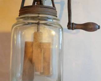 This is a glass butter churn featuring a metal gear mechanism and wooden paddles inside. The churn has a hand crank for manual operation and a metal lid. The construction is indicative of early to mid-20th century household butter churns.