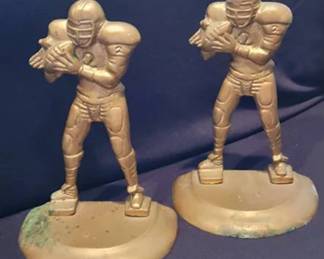 This pair of cast metal bookends features football players in uniform, each holding a football. Both figures have the number 2 on their jerseys and stand on curved bases. Visible surface patina and some discoloration are present. Approximate height is 8 inches based on scale reference.