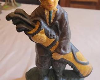 Cast iron doorstop depicting a golfer in period attire holding a golf bag, painted in brown, green, and yellow. Measures approximately 8 inches in height. Features felt pads on the base to protect surfaces. Suitable as a decorative doorstop or collectible figure.