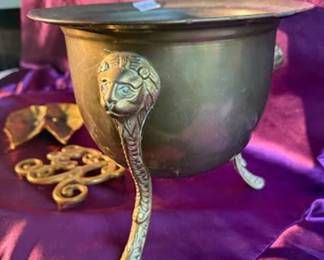 Brass lion-head footed pot, square-based brass candlesticks, a brass decorative leaf dish, and a brass monogram ornament. The brass pot has three detailed lion-head legs for support. 