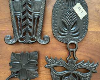 Cast iron trivets with various decorative motifs, including floral and pineapple designs. Each trivet has hanging holes and raised patterns. Maker's markings visible on the reverse sides.