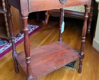 Rectangular wood side table featuring a single drawer with a decorative pull and a lower shelf. It stands on four turned legs. The table surface shows visible scratches and wear. The drawer is constructed with metal fasteners.
