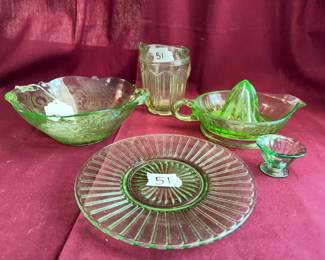 Green Depression glass tableware, including a ribbed plate, patterned bowl, handled citrus reamer, small footed bowl, and paneled glass cup. Each piece shows classic green glass coloring and period design elements. 