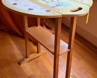 Wooden drop-leaf table featuring a painted tray top with a yellow house and tree motif, heart accents, and side handles. Dimensions: 24.5"H x 21"D x 29"W. Lower shelf included for stability and storage. 