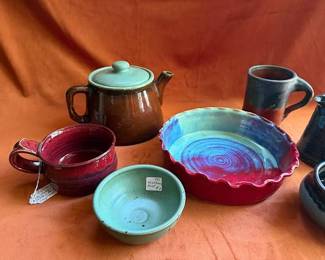 Studio pottery including a brown teapot with a pale lid, red and green mugs, a turquoise bowl, a blue and red fluted dish, and multiple blue glazed pitchers and bowls. Several pieces are signed, including by artists Bolick and Harwood.