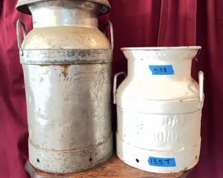 Metal dairy milk cans with side handles and embossed logos. One can is marked 'Engle's Dairy,' unpainted, approximately 19 inches tall. The other is painted white with 'Virginia Dairy Richmond VA,' labeled '13.5 T' in marker.
