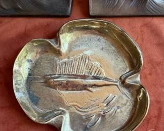 Brass sailfish-form bookends and a brass ashtray featuring a relief fish design. The bottom of the ashtray is marked with a round maker's-stamped logo and numerical code J-10. 