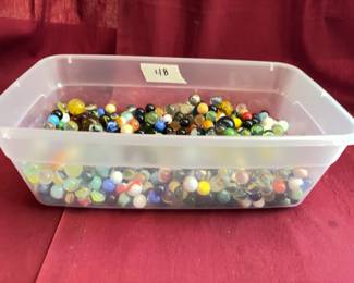 Glass marbles in various colors and designs, contained in a clear plastic bin. Suitable for collectors or general use. The lot includes an assortment of shooter and standard-size marbles.