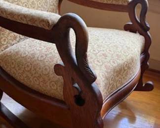 Upholstered goose head rocking chair featuring a patterned fabric with a floral motif. The high backrest and seat are framed by carved wood, including detailed cresting and scrolling arms. The wood has a rich, warm finish.