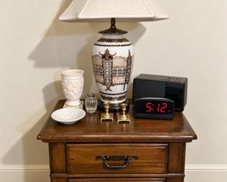 Classic two-drawer wooden nightstand featuring a warm finish, decorative hardware, and traditional styling. Also pictured are an ornate ceramic table lamp with shade, digital alarm clock, radio, brass candlesticks, and coordinating white ceramic and glass accessories—perfect for a bedroom, guest room, or sitting area.