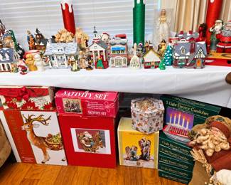A large selection of Christmas décor and collectibles, including lighted village houses and churches, Santa and angel figures, ceramic trees, holiday bells, nativity pieces, oversized glitter candles, mugs, and other festive accents. Boxed items include nativity sets, a decorative holiday reindeer, collectible figures, and multiple five-light electric candoliers. Plenty of treasures for holiday decorators and collectors!