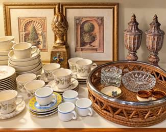 Elegant collection of vintage china, home décor, and serving pieces! This display includes floral teacups and saucers, stacked dishes, espresso cups, crystal bowls and coasters, a large woven serving basket, coordinating framed topiary prints, an ornate gold finial, and a beautiful pair of decorative lidded urns. Perfect for entertaining or adding traditional charm to your home!