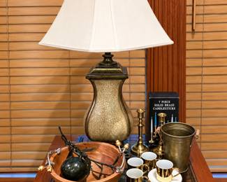 Worked for 15s  An elegant assortment of home décor and entertaining pieces, including a decorative table lamp, solid-brass candlestick set, brass ice bucket, gold-trimmed cups and saucers, a large wooden serving bowl, sculptural metal accents, and other unique decorative treasures.