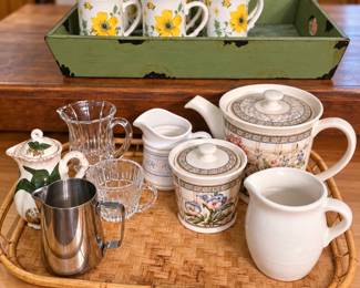 Charming assortment of kitchen and serving pieces, including a floral teapot with matching covered sugar bowl, ceramic and glass creamers, crystal cups, a stainless-steel pitcher, and a classic white pitcher—all displayed on a woven tray. Also featured is a matching set of cheerful yellow-floral mugs in a rustic green wooden tray. Perfect for entertaining, collecting, or everyday use!