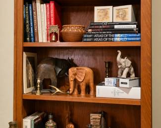A wonderful collection for book lovers and décor enthusiasts! This display includes a handsome wooden bookcase filled with classic literature, encyclopedias, art books, atlases, and other collectible titles. Also featured are carved wooden and metal elephants, a large elephant figurine, pottery, decorative vases, brass candlesticks, a magnifying glass, framed art, and additional unique accents.