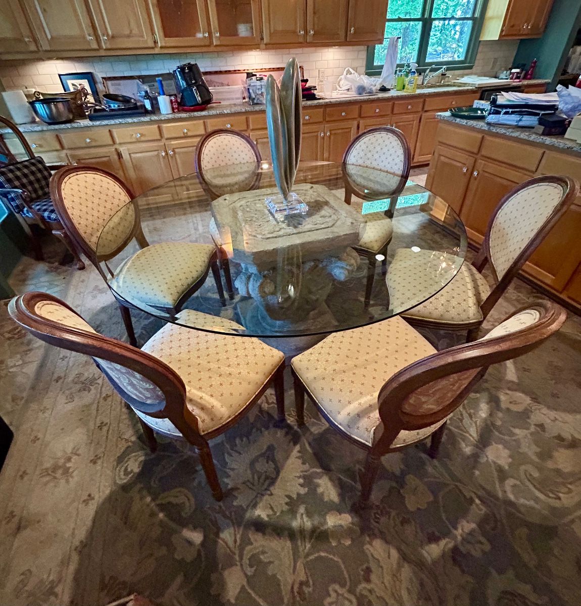 Round glass-top dining table with an ornate carved stone pedestal base and six upholstered oval-back chairs.