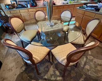 Round glass-top dining table with an ornate carved stone pedestal base and six upholstered oval-back chairs.