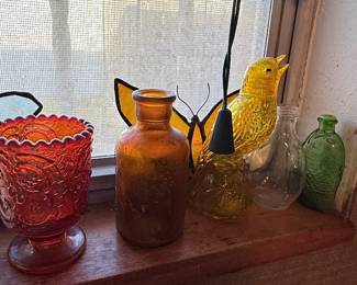 Assortment of decorative glass including a ruby red pressed-glass pedestal vase, amber bottle, yellow bird figurine, and green and clear vintage bottles.
