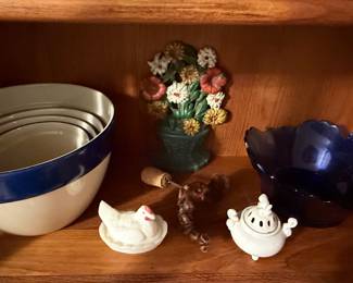 Assortment of vintage kitchen and decorative pieces including nested enamel mixing bowls with cobalt blue rims, cobalt blue glass serving bowl, floral wall décor, ceramic hen dish, and small decorative figurines.