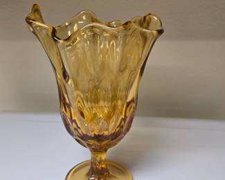 Bring vibrant mid-century warmth to your home with this stunning Fenton art glass pedestal vase. Rendered in a rich Colonial Amber / Honey gold colorway, this collectible features a classic geometric thumbprint pattern pressed into the lower half of the bowl. The top features a dramatic, hand-stretched "handkerchief" or "swung" rim that flares out into beautiful, fluid ruffles. Supported by an elegant panel-cut pedestal stem and weighted round base. A spectacular standalone statement piece or addition to any retro art glass collection.