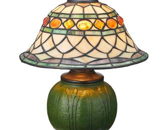 3395 An Arts & Crafts style leaded glass lamp Early 21st century The single light lamp with a composite base in the style of a Grueby Faience pottery vessel, surmounted by a leaded glass shade, electrified Unmarked Height of lamp with harp 12 1/2 in., diameter 6 in.; height of shade 5 1/8 in., diameter 10 3/8 in. Estimate: $300 - $500