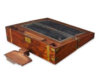3387 An English walnut lap desk and brass inkwell 19th/early 20th century The lid opening to reveal a gilt-stamped black leather writing surface with storage underneath, a pen rest, and other storage components, with an Art Nouveau inkwell 2 pieces Height of slope 7 in., width 20 in., depth 10 in.; height of inkwell 1 7/8 in., width 7 7/8 in., depth 6 1/4 in. Estimate: $300 - $500