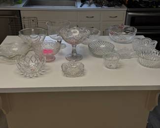 a collection of clear pressed glassware displayed on a white counter, featuring footed bowls, a large pedestal centerpiece bowl, and various scalloped serving dishes
