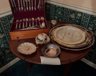 round wooden accent table holding a flatware storage box of Tara sterling silver, silverplate serving trays, a divided glass dish, and a covered serving piece