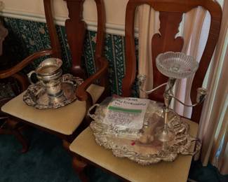 Queen Anne style dining chairs holding silverplate serving pieces, including an ornate tray, a pitcher, a candelabra, and packaged flatware