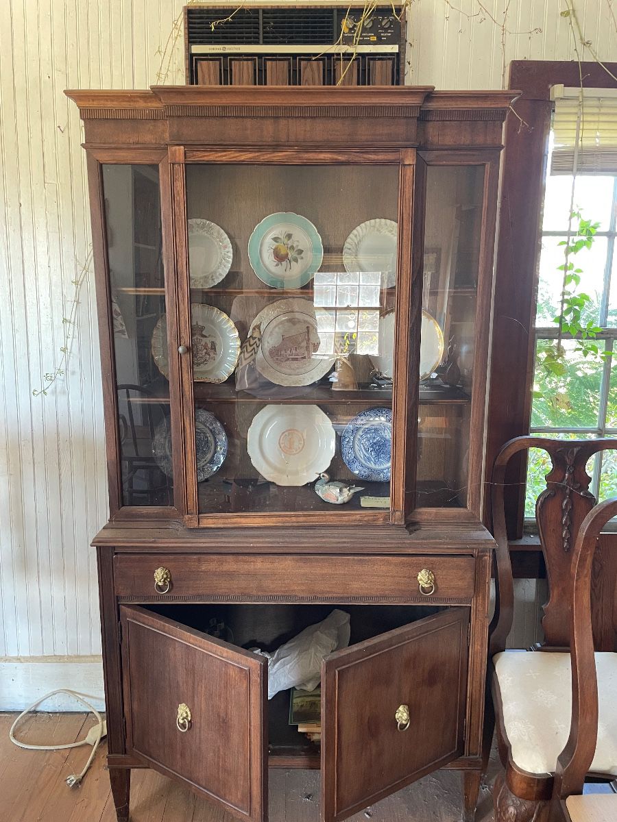 This is a vintage mahogany china cabinet featuring a glazed upper display section with glass doors and side panels enclosing wooden shelves currently holding decorative plates. The lower section consists of a long drawer over a double-door cabinet base, accented with brass ring pull hardware.