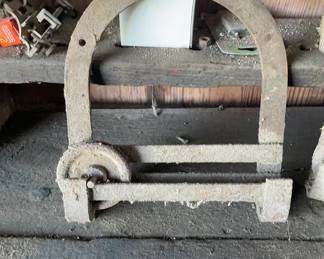 1880-1920 Antique Barn Door Roller This is a heavy cast iron antique barn door roller featuring an arched bracket with mounting bolt holes and a single grooved sliding wheel mechanism. The surface is heavily weathered, coated in a layer of dust and rust patina typical of prolonged barn storage.