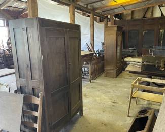 Armoire 1880-1920 A large wooden freestanding wardrobe with two front doors, panel details, and a dark finish, situated in a cluttered rustic workshop setting.