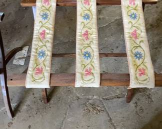 One of two Vintage luggage racks 1950s This folding wooden luggage rack features three cream-colored fabric straps adorned with an embroidered floral vine pattern featuring pink and blue flowers. The wooden frame is constructed with foldable X-legs and horizontal stretchers supporting the straps.