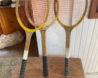 1950-1970 Three vintage wooden tennis racquets feature laminated wood frames with orange strung nylon nets, leather grips, and painted accents in yellow and white along the throats and heads.