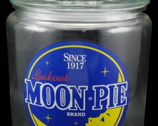 189 - Glass Moon Pie Store Jar 10.5"x7" (Description): Glass Moon Pie store jar featuring the  classic Lookout Moon Pie Brand logo with  "Since 1917" text. The clear glass jar has a  round shape with a fitted glass lid topped by  a knob finial. The front displays the iconic blue and yellow Moon Pie logo with the  smiling crescent moon design. Measures 10  1/2" x 7".