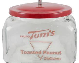 114 - Tom's Peanuts Glass Store Jar 8.5"x8"x8" (Description): Tom's Toasted Peanuts glass store jar  featuring the classic "enjoy Tom's" and  "Delicious" advertising graphics in red. The  clear glass jar has a rounded square shape  and is topped with a red metal lid with a chrome knob finial. This type countertop  display jar was commonly found in general  stores and service stations. Measures 8.5" x  8" x 8".