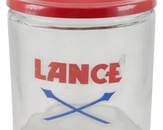 110 - Lance Glass Store Jar w/ Red Metal Lid (Description): Lance glass store jar featuring the iconic  red Lance logo with blue crossed arrows  design. The cylindrical clear glass jar is  topped with a red metal lid with a chrome  knob finial. This counter display jar was commonly used in general stores and service  stations to display Lance snack products.
