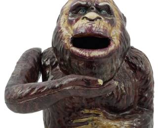 84 - Cast Iron Bigfoot Mechanical Bank (Description): Cast iron Bigfoot bank featuring a painted  bust of the legendary creature with an open  mouth, likely serving as the coin slot. The  bank depicts Bigfoot in a classic pose with  arms positioned across the chest. The piece shows detailed hand-painted coloring with  brown and tan tones highlighting the fur and  facial features, including expressive  blue-green eyes.