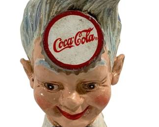 1521 - Cast Iron Coca-Cola Bank 8.5"x4"x5" (Description): Cast iron Coca-Cola advertising bank in the  form of a whimsical sprite or elf figure with  flowing gray hair and pointed ears. The  character features a Coca-Cola bottle cap  embedded in the forehead, rosy cheeks, red lips, and wears a white collar with a blue  bow tie. Hand-painted details throughout.  Measures 8 1/2" x 4" x 5".
