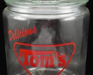 191 - Glass Tom's Roasted Peanuts Store Jar 10.5x7" (Description): Glass Tom's Peanuts store jar featuring red  printed advertising that reads "Delicious  Tom's Roasted Peanuts An American Tradition"  with the iconic Tom's shield logo. This  classic country store display jar has a round body with a glass lid topped with a ball  finial knob. Measures 10 1/2" x 7".
