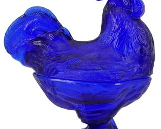 135 - Cobalt Glass Rooster Covered Dish 9" (Description): A striking cobalt blue glass rooster on  covered dish, standing 9 inches tall. The  pressed glass displays intricate feather  detailing throughout the rooster's body. The  deep cobalt blue coloring is consistent throughout, creating a bold visual statement  characteristic of vintage American pressed  glass collectibles.