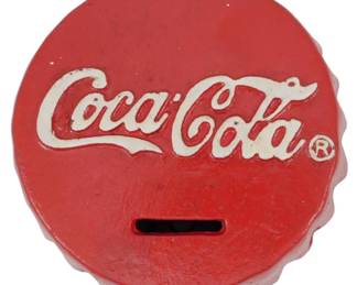 102 - Cast Iron Coca-Cola Bottlecap Bank 1.5"x5" (Description): Cast iron Coca-Cola bottlecap bank featuring  the classic red and white color scheme with  the iconic Coca-Cola script logo and  registered trademark symbol. The bank is  designed in the shape of a crimped bottle cap with a coin slot in the center. Measures  1.5" x 5".