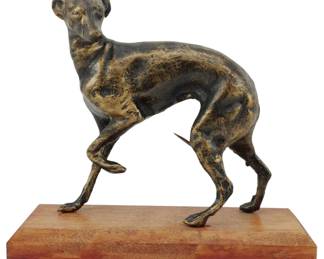 93 - Metal Greyhound Figure on Wooden Stand 6.5" (Description): A metal figure of a greyhound or whippet  mounted on a rectangular wooden stand. The  sculpture depicts the elegant dog in a  characteristic standing pose with one front  leg raised. The metal has a dark patinated finish with areas of gold-toned wear showing  through, giving the piece an antique  appearance. The figure measures 6.5 inches.