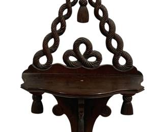 2095 - Wood Shelf 11"x19"x6.5" (Description): Wood shelf featuring an ornate carved rope  and tassel design. The shelf displays  decorative twisted rope motifs forming the  hanging element and knotwork details, with  carved tassels as embellishments. Measures 11" x 19" x 6.5".