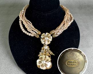 A mid-century vintage Miriam Haskell necklace of multi-strand baroque faux pearls with central clasp/pendant of two naturalistically carved mother-of-pearl petal flowers on gold-tone foliate frame accented with clear rhinestones. Clasp with raised oval Miriam Haskell logo. 15" length, including pendant, 20".