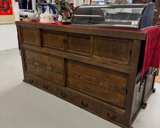 A large antique curved glass countertop display case, resting on the top section of a beautiful antique two-section Japanese Mizuya Tansu, a kitchen or tea room chest, with hand forged iron hardware. In two sections, the upper portion has two sets of sliding doors with storage space behind each, and five side-by-side drawers along the lower part. The bottom section has 2 sliding doors with spacious storage with shelving behind. The top cabinet measures 74”W x 20-1/4”D x 39”H and the lower cabinet measures 74”W x 20-1/4”D x 28”H.