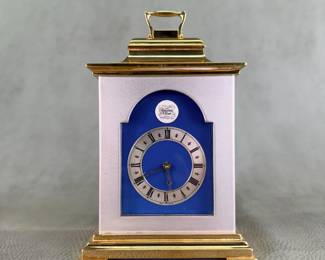 A vintage brass-gilded carriage clock / bracket clock with blue enamel framing, manufactured by the famous British clockmaking firm Thwaites & Reed. The clock runs; the face with stylized Roman numerals with star markers between them and elegant gold-toned openwork hands. A vibrant royal blue enamel arched backdrop surrounds the dial, and it is marked with the brand crest at the 12 o'clock position and "Made in England" subtly at the bottom. Comes with original paperwork from Thwaites & Reed Limited.